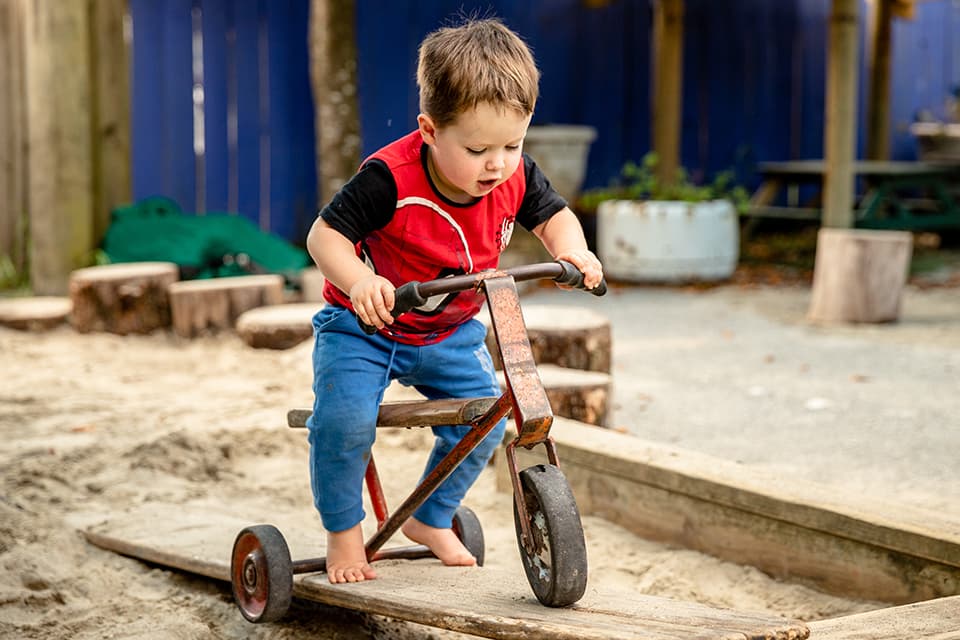 Young boy riding on tricycle over wooden plank in sandpit.