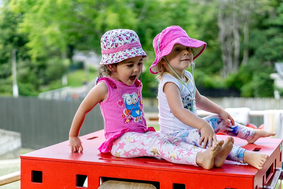 Two girls smiling in sun hats sitting on outdoor box in daycare glen eden.