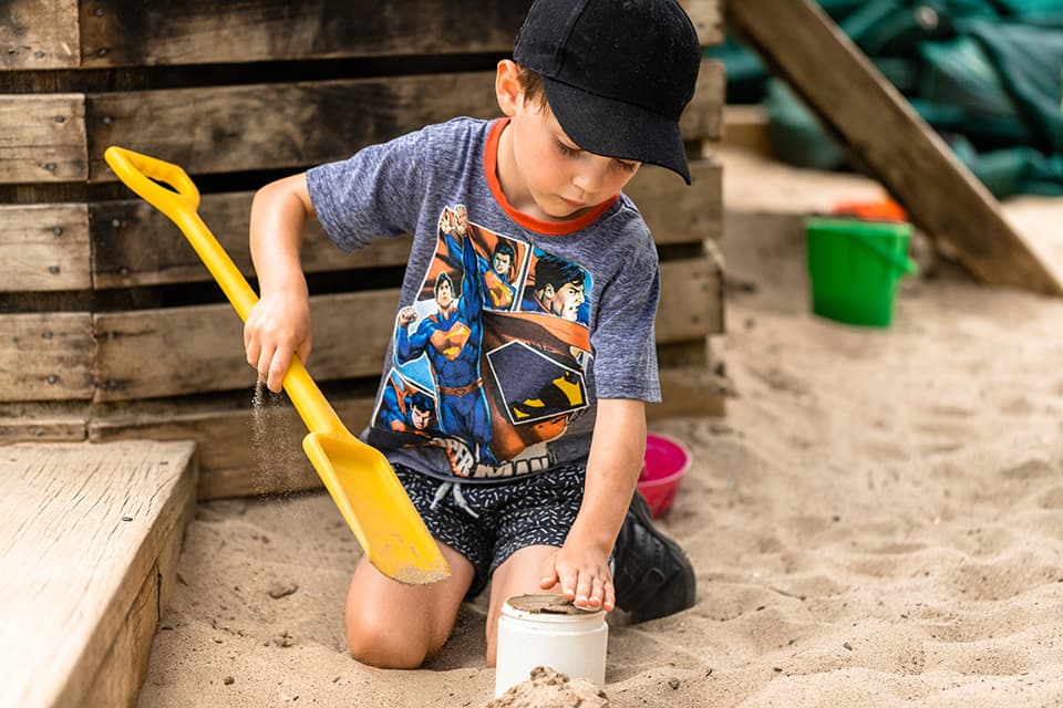 Young boy in hat filling up bucket with sand in sandpit.