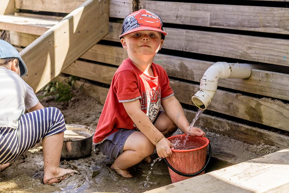 Young boy in hat filling up bucket with water in sandpit.